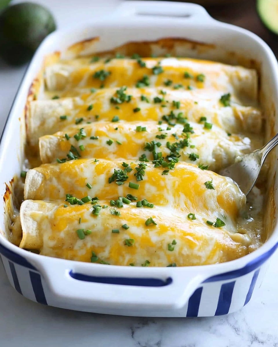 Smothered Green Chile Chicken Burritos, green chile chicken burritos, Mexican chicken burritos, creamy green chile sauce, flavorful burrito recipes The dish shows a white baking dish with blue stripes on the outside, filled with five rolled enchiladas covered in melted cheese. Each enchilada has a soft, light yellow tortilla base, covered fully with thick, creamy, white sauce and topped with a golden-yellow melted cheese layer that stretches slightly as one enchilada is lifted by a spatula. Small green chopped chives are sprinkled over the cheese, adding a fresh color contrast. The background is a white marbled surface with blurred avocado halves and a small bowl visible. The