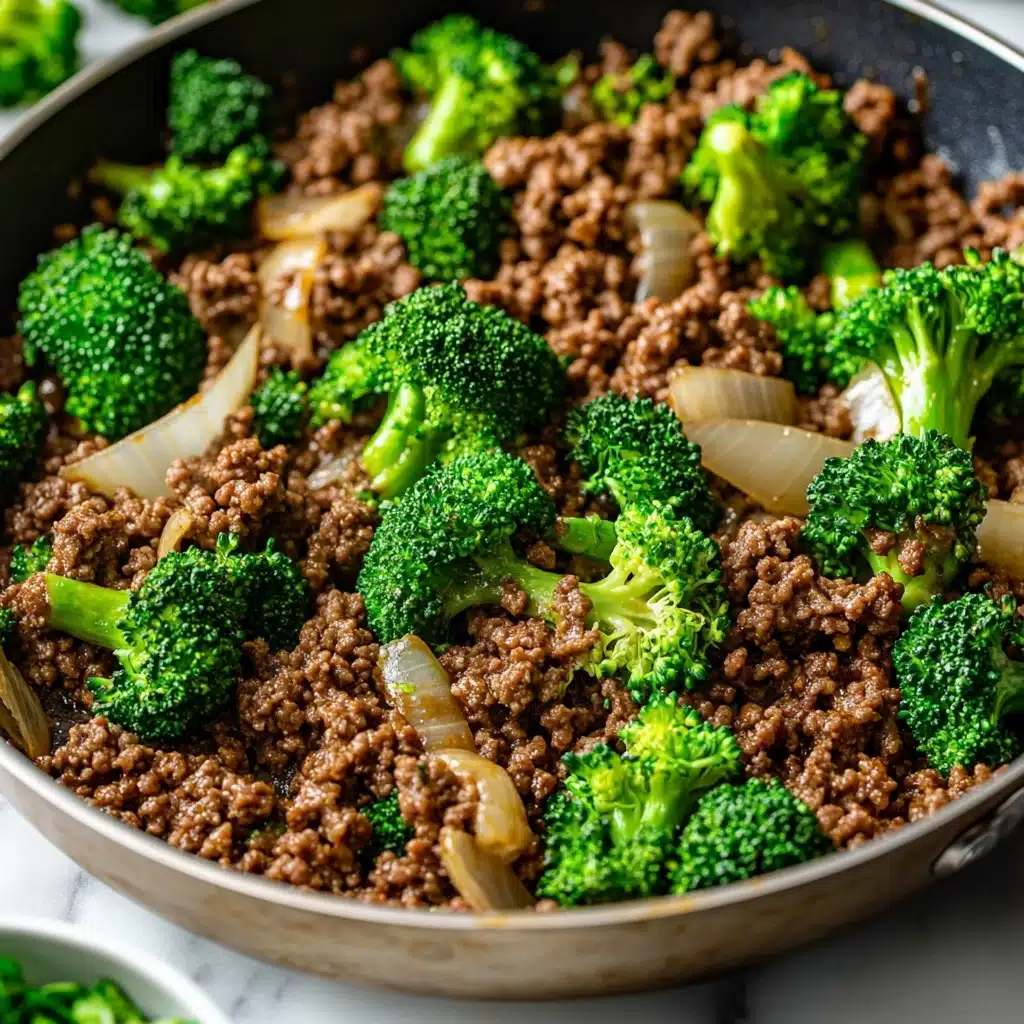 Ground Beef and Broccoli Stir Fry, quick beef stir fry, healthy broccoli dinner, easy weeknight stir-fry, family-friendly beef and broccoli A close-up view of a pan filled with cooked ground beef that is brown and crumbly as the base layer, mixed with pieces of cooked white onion that are slightly translucent and scattered evenly throughout. On top, bright green broccoli florets with a fresh, crisp texture are mixed in, adding a contrasting color and a fresh look. The pan is placed on a white marbled surface, and a small part of a small bowl with green sliced ingredients is visible near the bottom left corner. The overall scene captures a warm and hearty cooked dish, with a mix of textures from soft beef, tender onions, and fresh broccoli