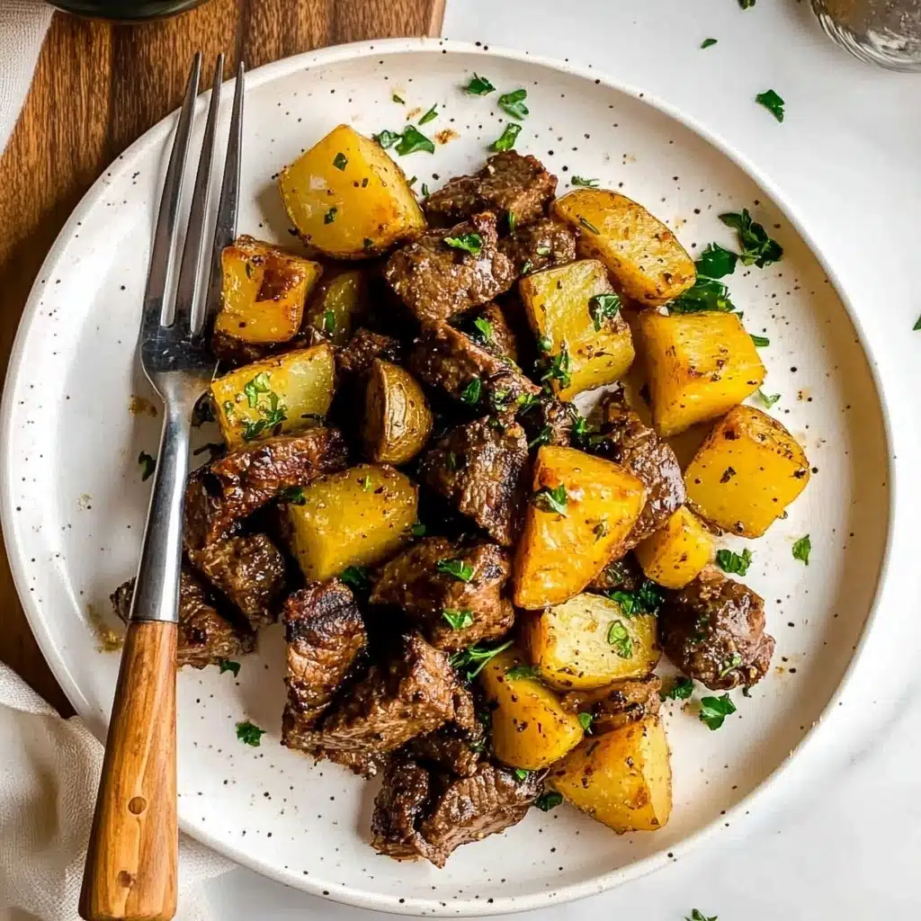 Garlic Butter Steak Bites and Potatoes, Steak Bites and Potatoes, Easy Steak and Potato Dinner, One-Skillet Steak and Potatoes, Quick Weeknight Steak Dinner A white round plate holds a simple meal with two main layers: the first layer is golden yellow potato chunks with a slightly crispy texture, scattered around the plate, some showing slight browning edges; the second layer is browned beef pieces, tender and juicy looking, mixed evenly with the potatoes and sprinkled with fresh green herbs on top for color contrast. A fork and knife with wooden handles are placed on the left side of the plate, resting on a white marbled surface. Shadows and light suggest natural daylight.