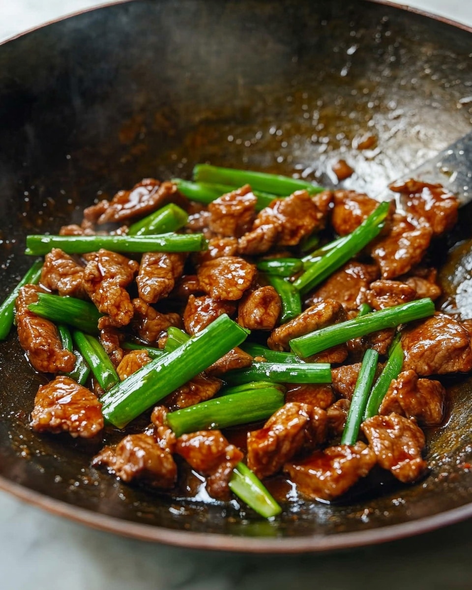 Mongolian Chicken, Mongolian Chicken recipe, Chinese chicken dishes, quick Asian dinner, crispy chicken stir-fry Inside a dark metal wok on a white marbled surface, small pieces of browned meat are mixed with bright green scallions. The meat looks shiny and cooked in a thick brown sauce that pools slightly at the bottom. The scallions are cut into long thin pieces and scattered evenly on top, adding a fresh contrast in color and texture. The wok shows signs of use, with some light marks and a textured surface inside, and a metal spatula edge is visible at the top right corner.