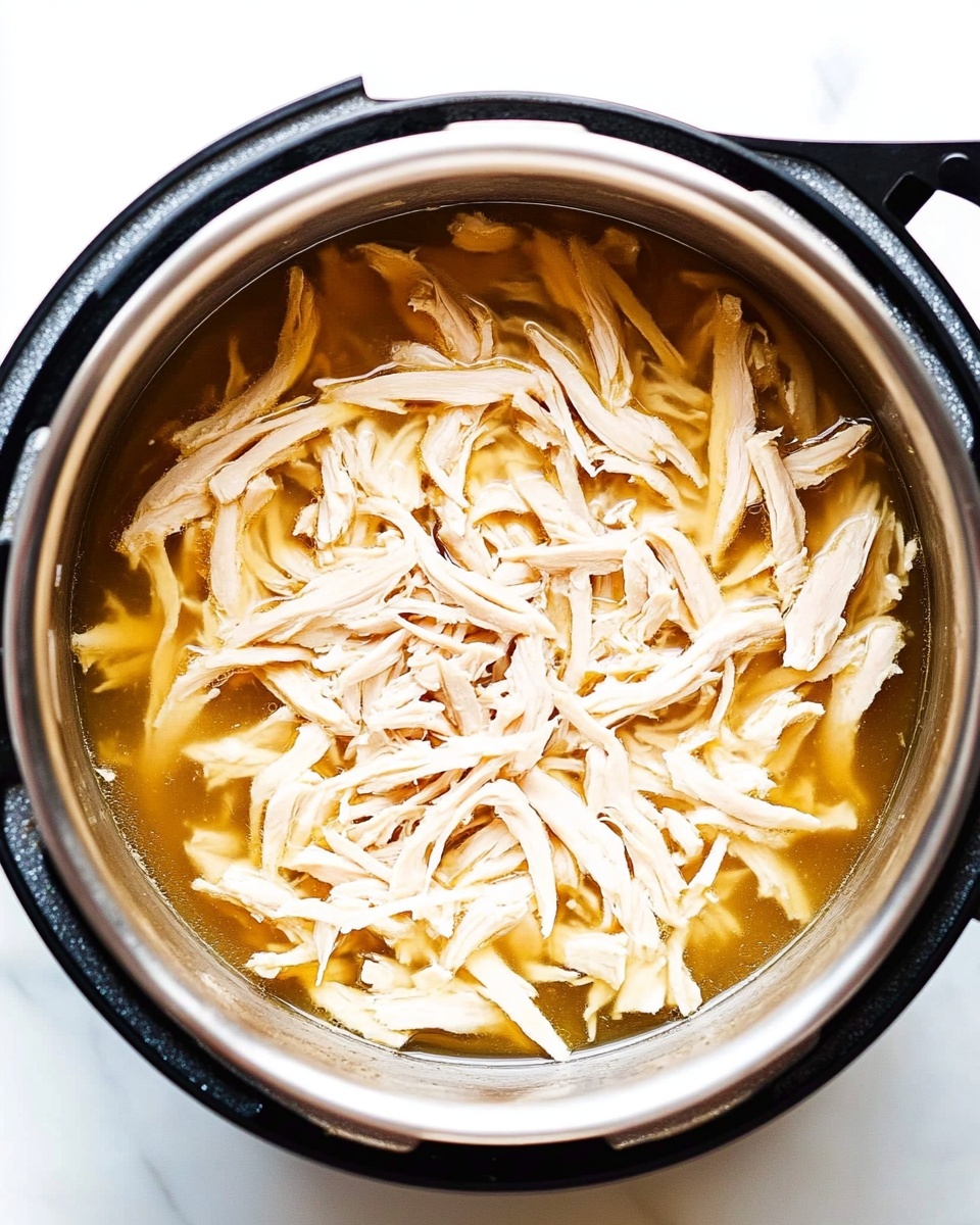 Instant Pot Shredded Chicken, quick shredded chicken, easy Instant Pot chicken, healthy shredded chicken recipe, versatile chicken for meal prep The image shows a close-up top view of shredded light beige chicken resting in a shallow layer of golden-brown broth inside a shiny silver metal pot. The chicken pieces are long, thin, and stringy, scattered unevenly throughout the pot. The pot has a black outer rim and handles, and it sits on a bright white marbled surface. The light reflects softly off the broth and the moist chicken, creating a fresh and clean look.