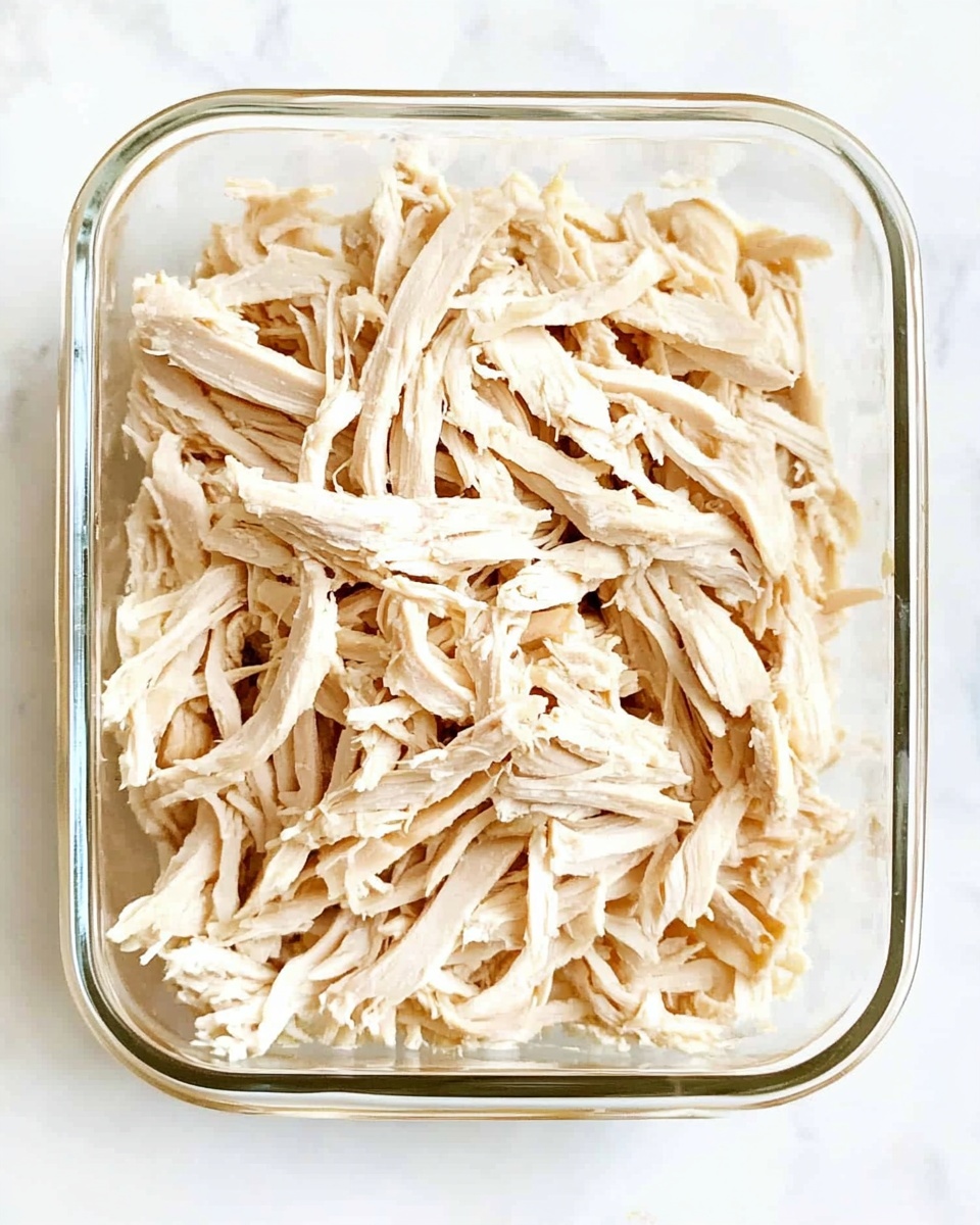 Instant Pot Shredded Chicken, quick shredded chicken, easy Instant Pot chicken, healthy shredded chicken recipe, versatile chicken for meal prep The image shows a clear glass rectangular container filled with shredded cooked chicken. The shredded chicken is light beige, with soft, fibrous texture pieces layered loosely to fill the container evenly from top to bottom. The chicken strands are thin and varied in size and shape, creating a natural, slightly uneven surface. The container sits on a white marbled surface.