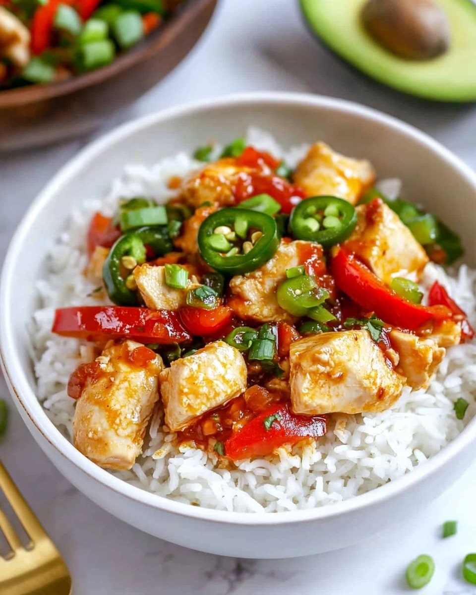 Sweet Chili Chicken Delight, sweet chili chicken, baked chicken with sweet chili sauce, easy chicken dinner, quick weeknight chicken meal A white bowl is filled with a base layer of fluffy white rice. On top of the rice, there are pieces of cooked chicken in a light golden color, mixed with bright red strips of bell pepper and dark green sliced jalapeños. The dish is garnished with chopped green onions and drizzled with a shiny red chili sauce. In the background, there is another similar white bowl and a wooden bowl with more chopped green onions placed on a white marbled surface. A gold fork and half an avocado are also seen nearby.