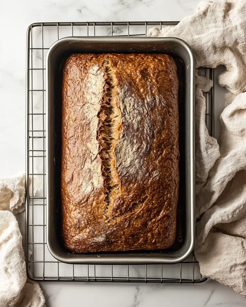 Simple Homemade Banana Bread, easy banana bread recipe, moist banana bread loaf, beginner banana bread, quick banana bread A rectangular loaf of brown bread with a cracked top surface rests in a silver metal loaf pan, placed on a metal cooling rack over a white marbled texture. The bread's crust is evenly baked with a golden brown color and some darker spots, showing a rough texture. Next to the pan, a beige fabric cloth is casually placed, adding a soft touch to the scene.