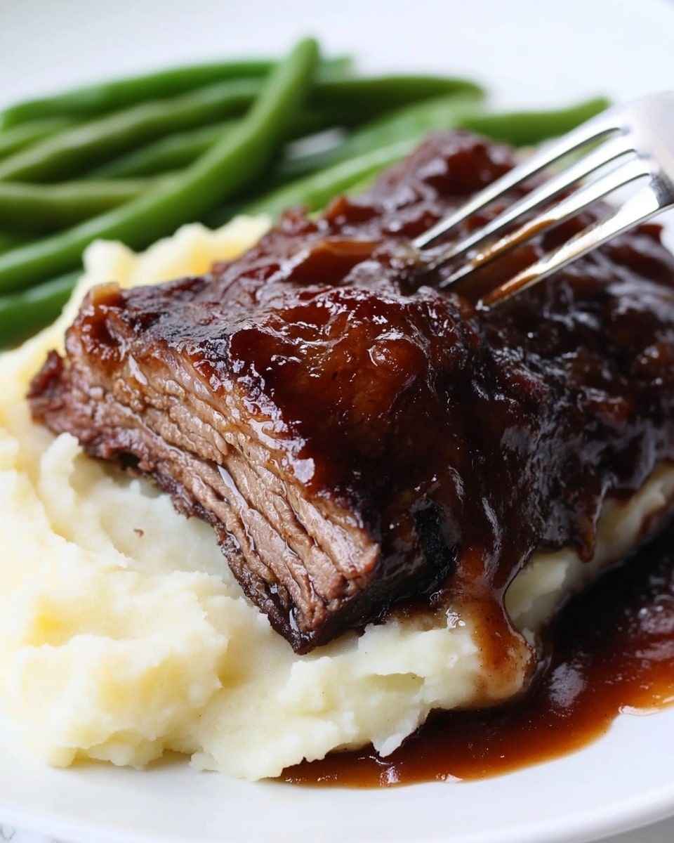 Instant Pot Beef Ribs, crispy broil finish, easy beef rib recipe, quick pressure cooker ribs, tender beef ribs The image shows a white plate with three food layers and a utensil in front. The bottom layer is mashed potatoes, which look smooth and creamy with a soft, fluffy texture, colored white to light yellow. On top of the mashed potatoes is a thick, dark brown barbecued meat piece with a shiny, caramelized sauce glaze and a slightly rough texture. To the right of the plate, green beans add a fresh and crisp green layer with a smooth surface. The background features a white marbled table surface and a blurred kitchen appliance, creating a cozy kitchen feel.