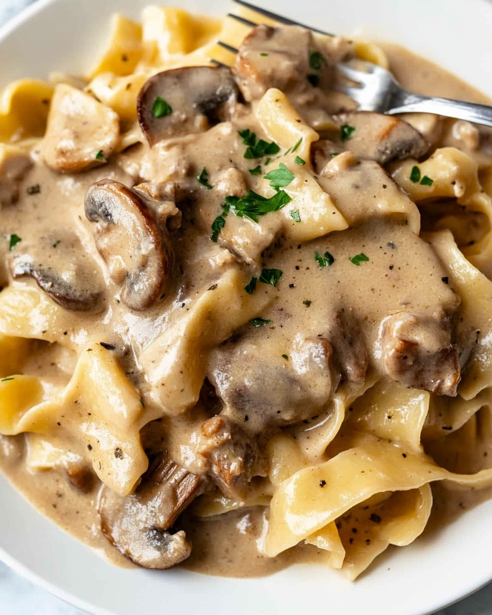 This image shows a white plate filled with wide, flat pasta noodles layered underneath and mixed with thick, creamy beige sauce that has visible specks of herbs. On top and throughout the pasta are pieces of tender-looking brown meat and sliced mushrooms, all coated evenly by the sauce. Small bits of chopped green parsley are sprinkled across the dish, adding a touch of color to the creamy textures. A silver fork rests at the top edge of the plate, partially stuck in the food. The plate is set on a white marbled surface. photo taken with an iphone --ar 4:5 --v 6.1