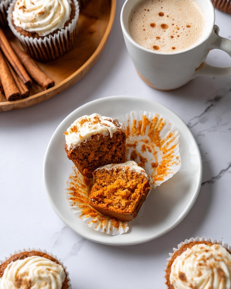 The image shows a white plate with an open muffin split into two halves revealing a moist, orange-brown crumb inside with some white cream cheese frosting on top and around the edges. The muffin liner is peeled back and lays crumpled on the plate, stained with orange crumbs. Surrounding the plate are whole muffins with swirled white and light brown cream cheese frosting in white liners. A ceramic cup filled with a foamy, light beige drink with small specks of cinnamon on top sits behind the plate. There is a wooden plate with cinnamon sticks in the upper left corner and the surface is a white marbled texture. photo taken with an iphone --ar 4:5 --v 7