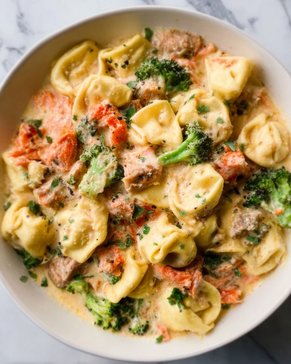 The image shows a close-up of a white plate filled with a creamy pasta dish, layered with tortellini that are pale yellow and slightly textured. The tortellini are mixed with pieces of green broccoli and orange-red chunks of salmon, all coated in a smooth, light cream sauce giving a glossy look. The ingredients are evenly spread, with the pasta forming the base layer and the broccoli and salmon scattered on top, creating a colorful and appetizing mix. The plate rests on a white marbled surface. photo taken with an iphone --ar 4:5 --v 7
