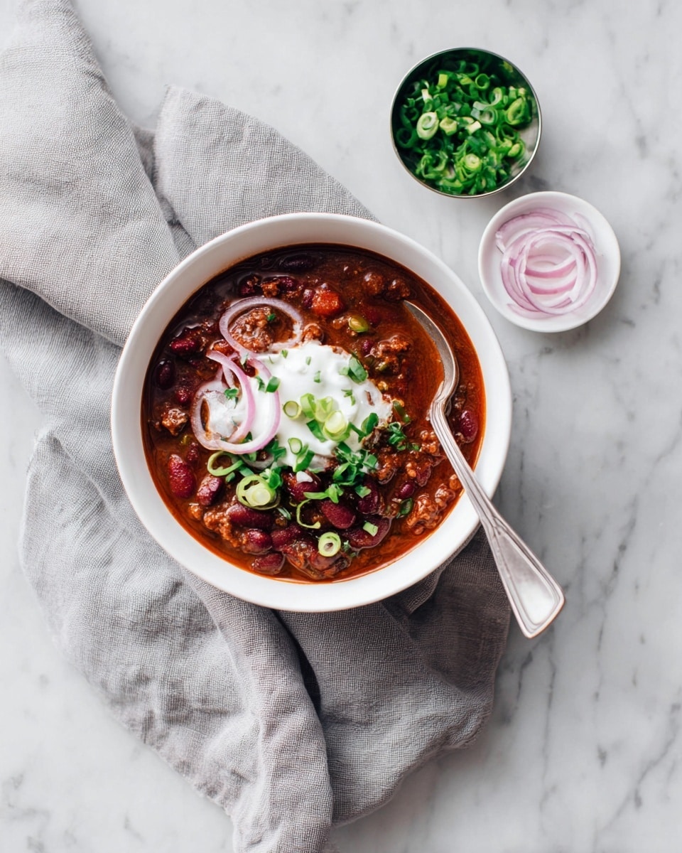 A white bowl filled with dark red chili made of beans and meat, topped with a dollop of white sour cream, a few thin light pink onion rings, and sprinkled with green chopped scallions. A silver spoon rests inside the bowl dipping into the chili. The bowl sits on a soft grey cloth on a white marbled surface. Next to the bowl, there is a small white dish with curled light pink onion slices and a small shiny metal container filled with fresh chopped green scallions. Photo taken with an iphone --ar 4:5 --v 7