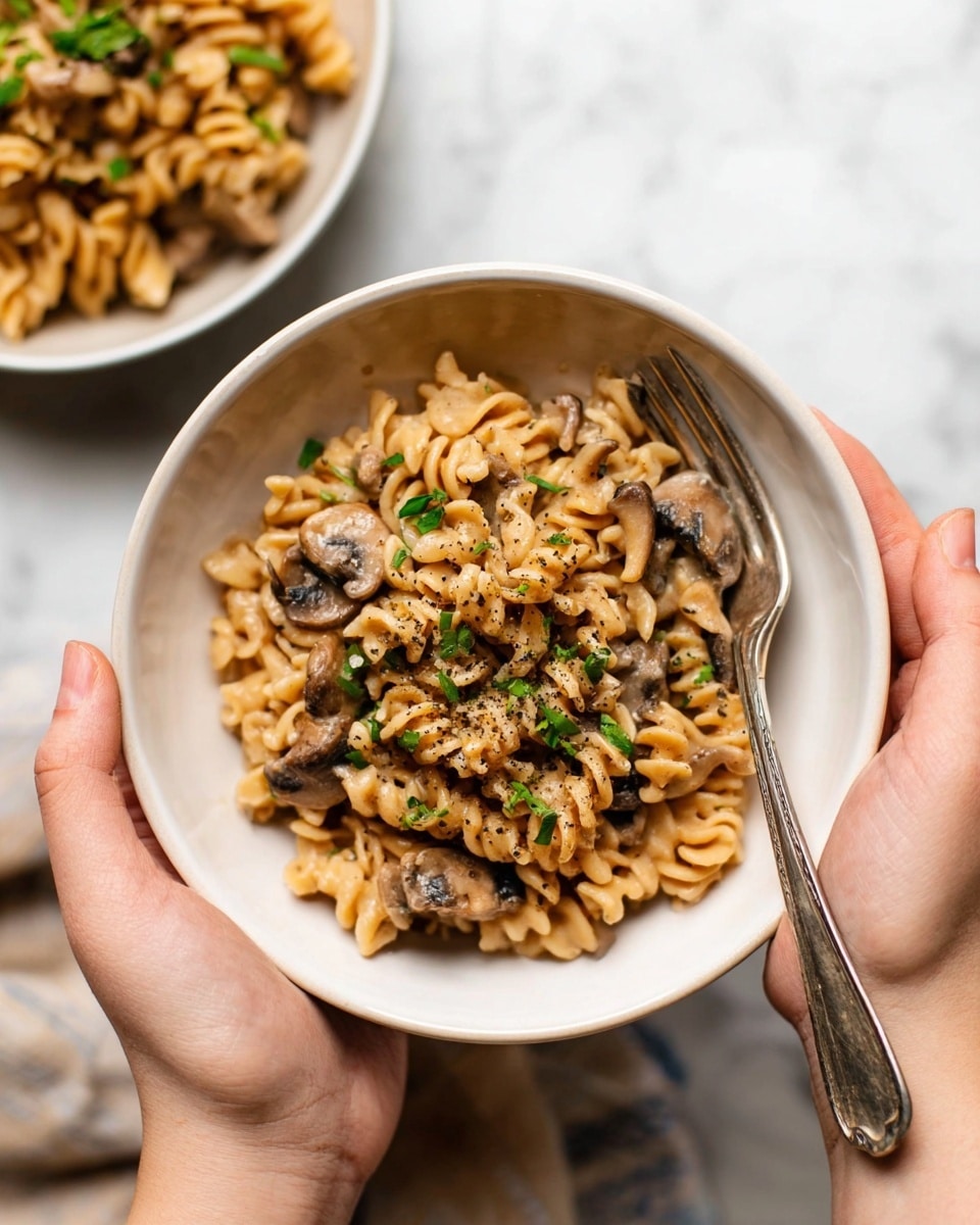 A close-up view inside a white pot filled with creamy, light brown sauce-covered rotini pasta mixed with sliced brown mushrooms, all coated evenly with small black pepper flakes scattered on top. A black spoon with a wooden handle is partially submerged in the pasta on the right side, stirring the mixture. The background shows a white marbled surface. Photo taken with an iphone --ar 4:5 --v 7