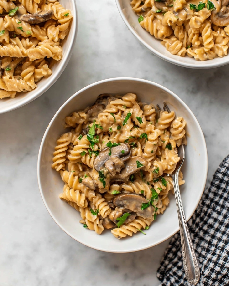The image shows a close-up of three white bowls filled with creamy mushroom pasta on a white marbled surface. The main bowl in the center contains spiral pasta coated in a thick light brown sauce, with visible slices of cooked mushrooms mixed throughout. The dish is garnished with small green parsley leaves sprinkled on top. A metal fork rests inside the main bowl on the right side. The other two bowls, partially visible at the top left and top right corners, also contain the same pasta dish. A checkered cloth is placed near the bottom right corner of the image. photo taken with an iphone --ar 4:5 --v 7