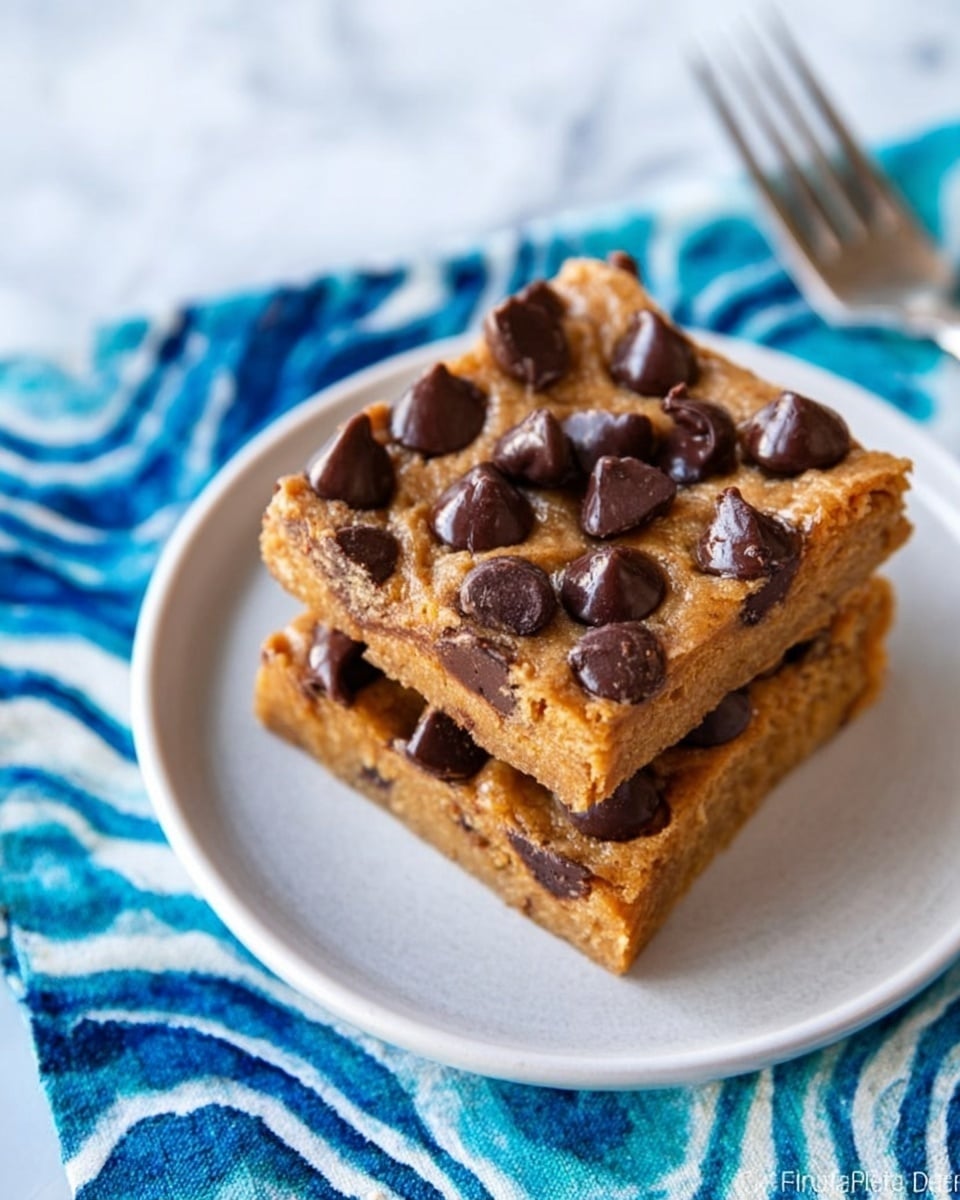 A stack of three thick, square pumpkin spice bars with a soft, crumbly texture sits on a small, wavy-edged dark blue plate. This blue plate is placed on a larger white plate, and both plates sit on a blue and white checkered cloth. In the background, there is an orange container with white