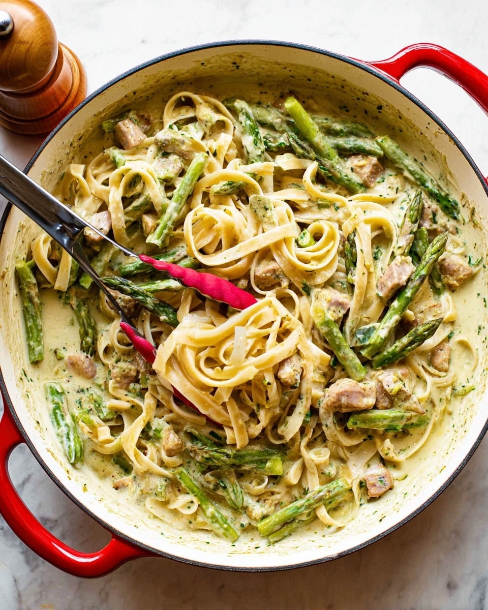 In a white round pan, there is a creamy pasta with three main layers: the bottom layer is a light green creamy sauce with some herbs mixed in, the middle layer consists of pale yellow, long, flat pasta noodles twisted around, and the top layer has small chunks of light brown cooked meat and bright green asparagus pieces scattered evenly. Red silicone-tipped tongs rest inside the pan holding a few pasta strands and asparagus, and the pan is placed on a white marbled surface with a wooden pepper grinder next to it. photo taken with an iphone --ar 4:5 --v 7