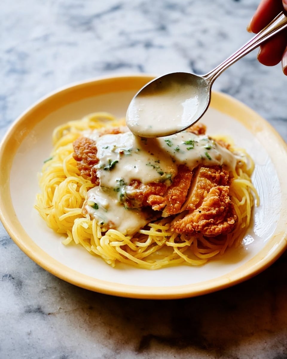 On a white plate, there is a small pile of thin yellow spaghetti noodles forming the base layer in the center. Next to the noodles on the right side, there is a piece of golden-brown fried chicken with a crispy texture. A silver spoon held by a woman's hand is pouring a creamy white sauce with green herb bits over both the noodles and the chicken. The background is a white marbled surface. photo taken with an iphone --ar 4:5 --v 7