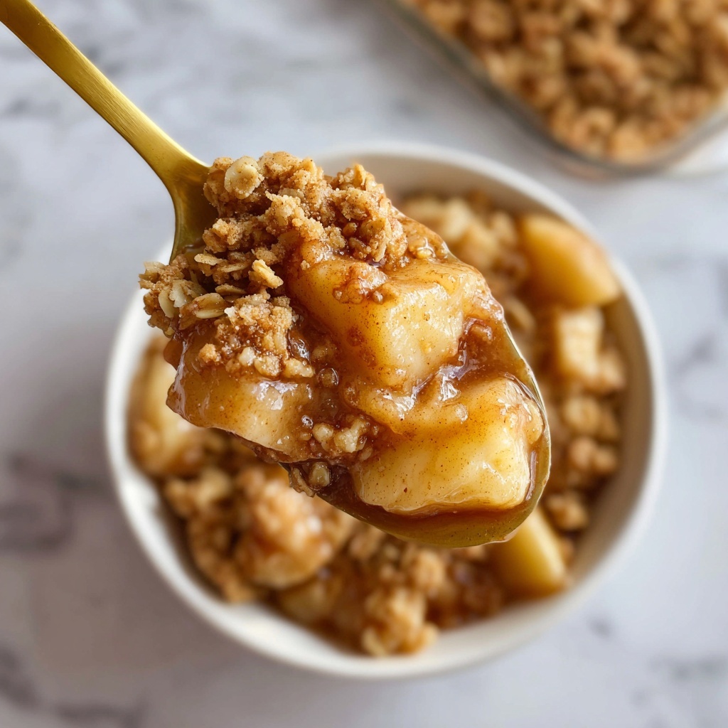 A close-up of a golden spoon holding a scoop of warm dessert made of soft, light brown apple pieces and a crumbly oat topping that looks crunchy and slightly sticky. The oat crumbs are light beige with a few cinnamon spots, scattered over the apples that are glossy and slightly translucent. The background is filled with more of the same dessert in a white bowl, showing layers of apple chunks and oat crumbs covered in syrupy sauce. The scene sits on a white marbled surface. photo taken with an iphone --ar 4:5 --v 7