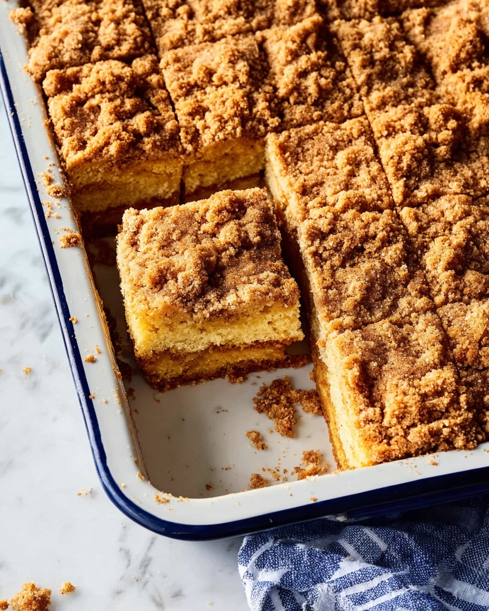 The image shows a white baking dish with a dark blue rim filled with crumb cake cut into squares. One square piece is lifted slightly, showing two thick layers of soft, light yellow cake with a crumbly golden brown topping that looks rough and crunchy. The cake's crumb topping covers the whole surface and some crumbs are scattered on the white marbled surface next to the dish. A piece of blue and white cloth is partially visible under the dish near the bottom right corner. Photo taken with an iphone --ar 4:5 --v 7