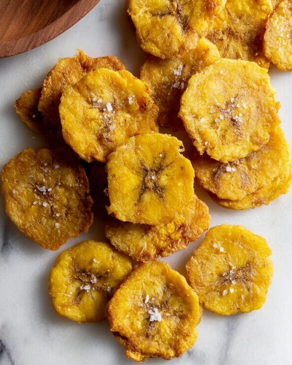 A close-up view shows a woman's hand holding a round, golden-brown fried plantain chip with a small bite taken out. The chip has a crispy texture with visible salt flakes on its surface. In the background, there is a white plate with more plantain chips piled on it and a small serving of white rice with beans on the side. A silver fork rests on the white marbled surface next to the plate. The colors are warm and natural, highlighting the crunchy texture of the chips. photo taken with an iphone --ar 4:5 --v 7