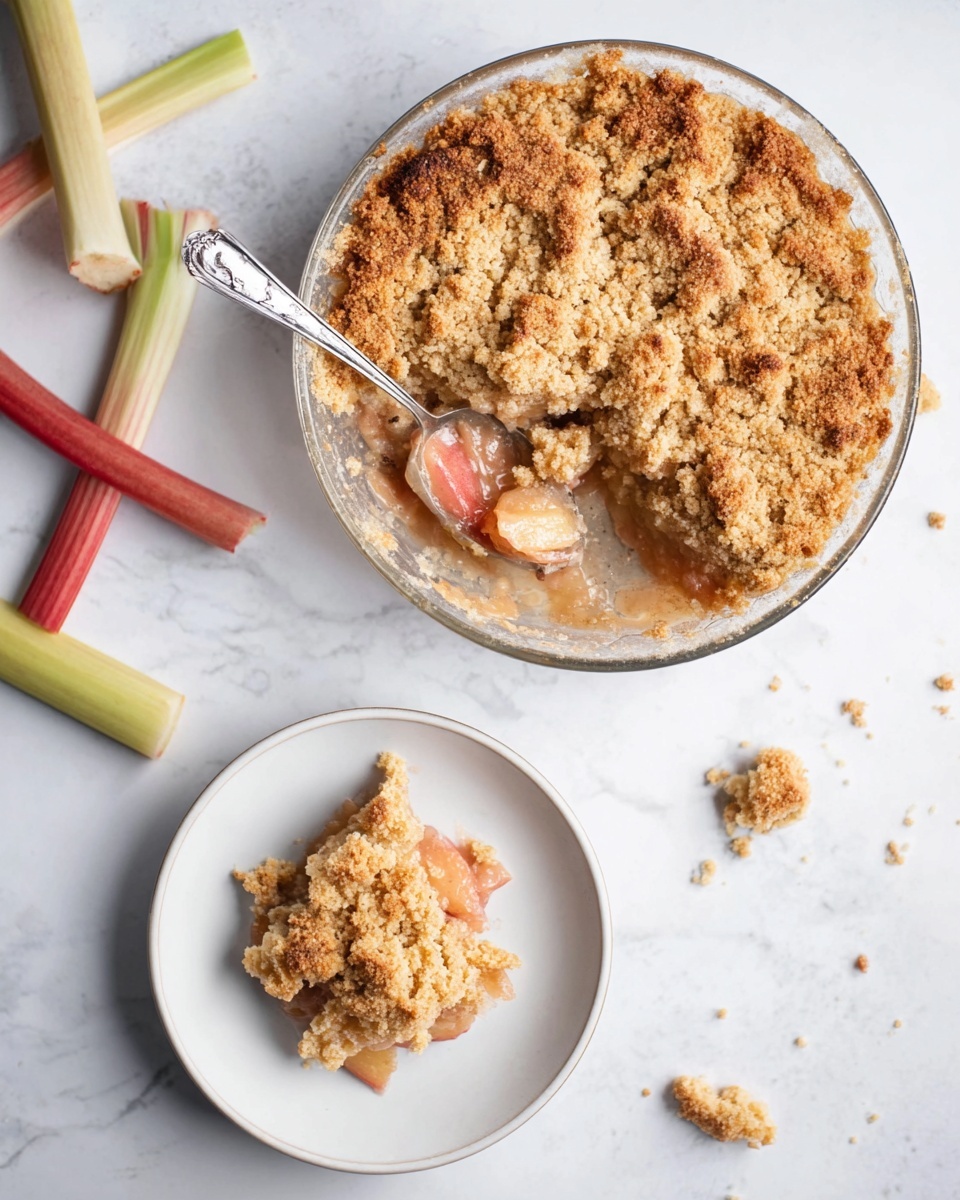 The image shows a round clear glass dish filled with a baked crumble that has a golden-brown, crumbly top layer evenly spread across. Inside, a peachy-pink fruit layer is partially visible where a spoon has scooped out a portion, revealing juicy soft fruit beneath the crumb layer. Next to it, a white plate holds one square piece of the crumble with visible light pink chunks under the topping. A shiny silver spoon rests on the plate, and scattered pieces of rhubarb stalks lie on the white marbled surface around the dishes. photo taken with an iphone --ar 4:5 --v 7