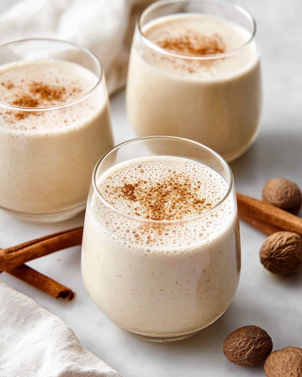 A close-up top view of a clear glass filled with a light beige foamy liquid being poured from a clear blender jar, showing bubbles on the surface of the drink. The glass is placed on a white marbled surface next to a round cork coaster. Photo taken with an iphone --ar 4:5 --v 7