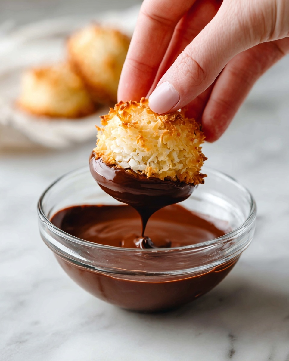 A woman's hand is holding a small round coconut macaron with a golden-brown crispy top layer and a rough shredded texture, dipping the bottom half into a smooth, dark brown melted chocolate layer inside a clear glass bowl. The bowl sits on a white marbled surface, and a second coconut macaron with a similar golden top is blurred in the background on the white marbled surface. photo taken with an iphone --ar 4:5 --v 7