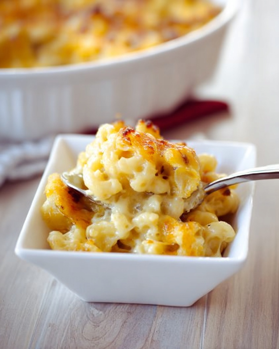 The image shows a small white square bowl filled with a portion of macaroni and cheese. The macaroni pasta is tube-shaped and covered in creamy yellow cheese sauce, with some parts browned from baking, making it look slightly crispy on top. A shiny metal spoon rests inside the bowl, holding a scoop of the cheesy pasta. In the background, a larger white round dish filled with more macaroni and cheese is slightly out of focus, sitting on a light wooden surface. Photo taken with an iphone --ar 4:5 --v 7