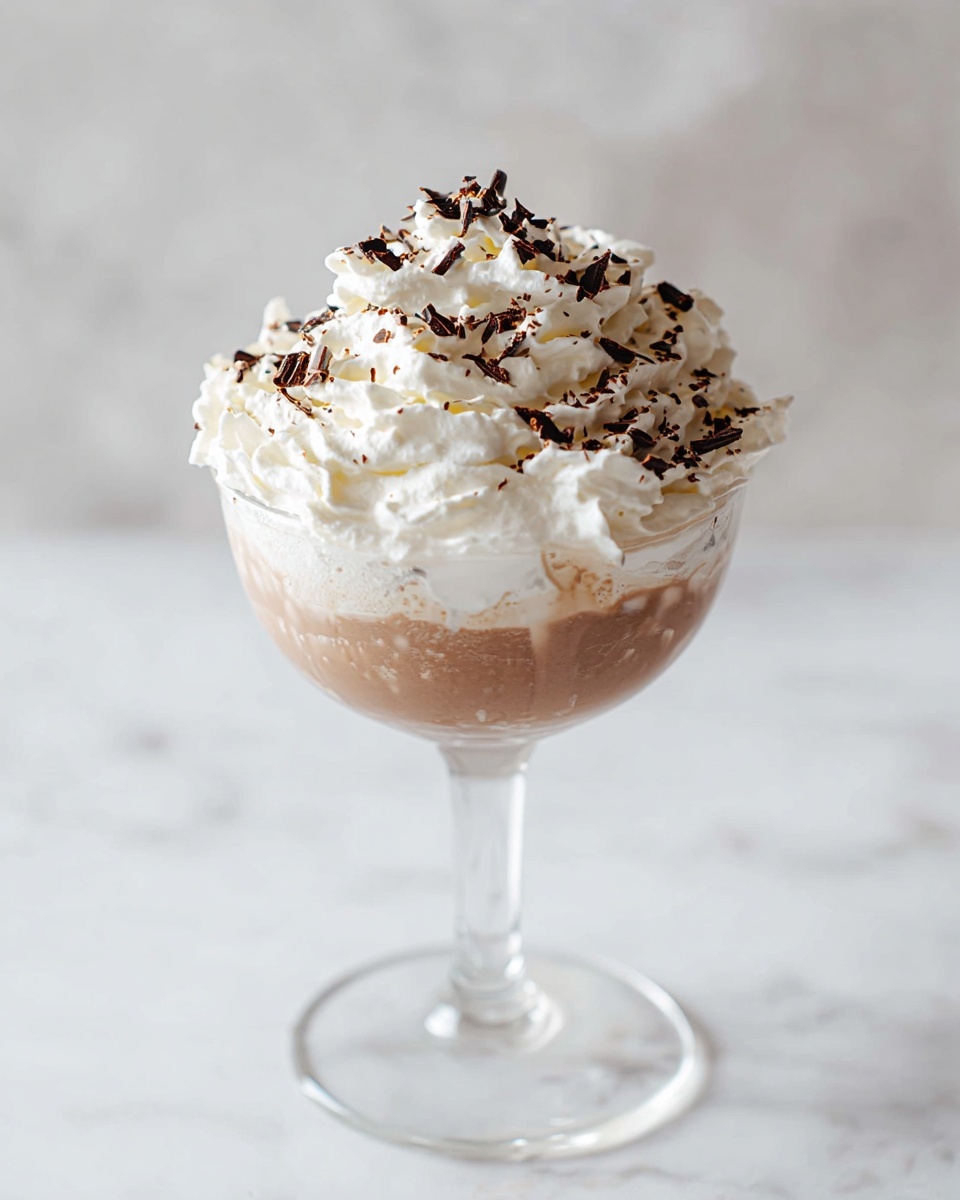 The image shows a clear glass dessert cup filled with two main layers. The bottom layer is thick and dark chocolate mousse with a smooth texture. On top of this, there is a thick, fluffy layer of white whipped cream, generously swirled in soft peaks covering the mousse. The whipped cream is sprinkled with small dark chocolate shavings, adding contrast and texture on the top. The cup is set against a white marbled background. photo taken with an iphone --ar 4:5 --v 7