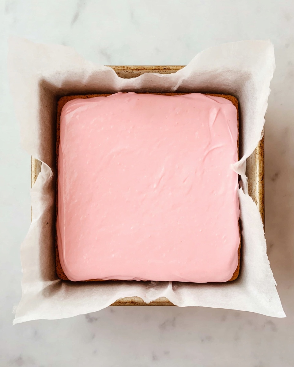 A square baking pan lined with white baking paper holds a two-layer dessert. The bottom layer is unseen but covered by a thick, smooth, pastel pink icing top layer that spreads evenly to the edges. The baking pan has a rustic look and sits on a white marbled surface, creating a clean and bright background. The pink layer looks soft and creamy with a shiny finish. photo taken with an iphone --ar 4:5 --v 7