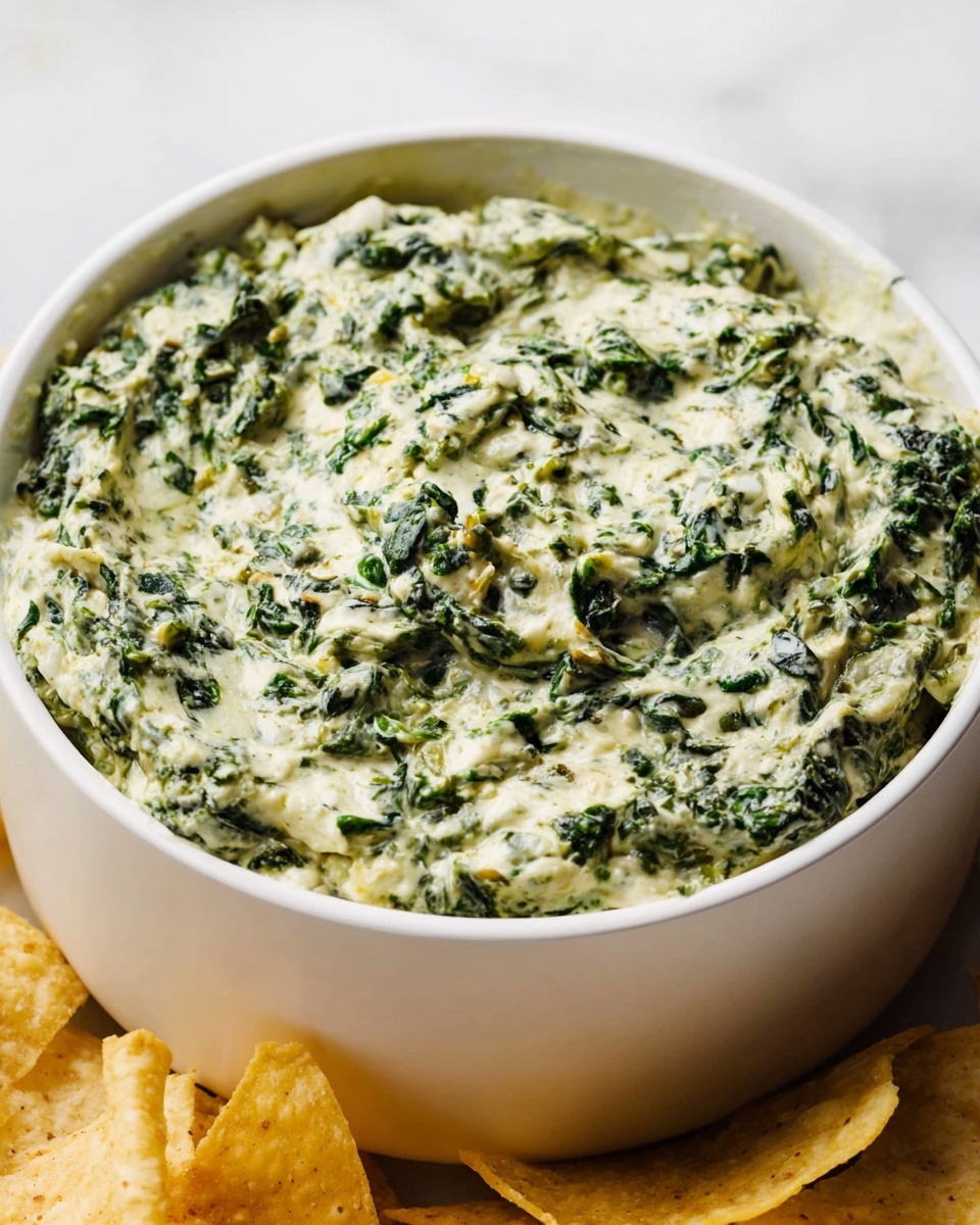 The image shows a white bowl filled with creamy spinach dip. The dip has two main colors: green from the spinach and light cream from the cheese sauce, mixed together evenly. The texture looks thick and slightly chunky because of the spinach bits, spread to the top edge of the bowl. The bowl sits on a white marbled surface, and there is a lid leaning on the back of the bowl. In the front, parts of some tortilla chips are visible, adding a light yellow color to the scene. photo taken with an iphone --ar 4:5 --v 7