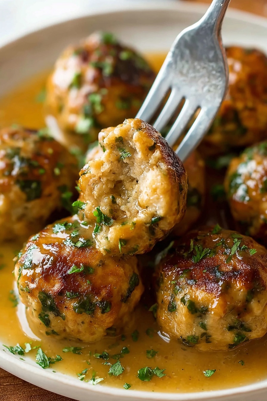 The image shows several round meatballs with a golden-brown crust and green herb bits scattered through them, resting in a light brown gravy. A silver fork holds one meatball with a bite taken out, showing a soft and moist inside with visible herbs. The meatballs are arranged closely on a white plate with some chopped green herbs sprinkled on top and the gravy pooling around them. The background is a white marbled texture photo taken with an iphone --ar 2:3 --v 7 - Stuffing Balls Holiday Side Dish, Easy Stuffing Balls Recipe, Festive Stuffing Bites, Cozy Holiday Side Dishes, Savory Stuffing Snacks
