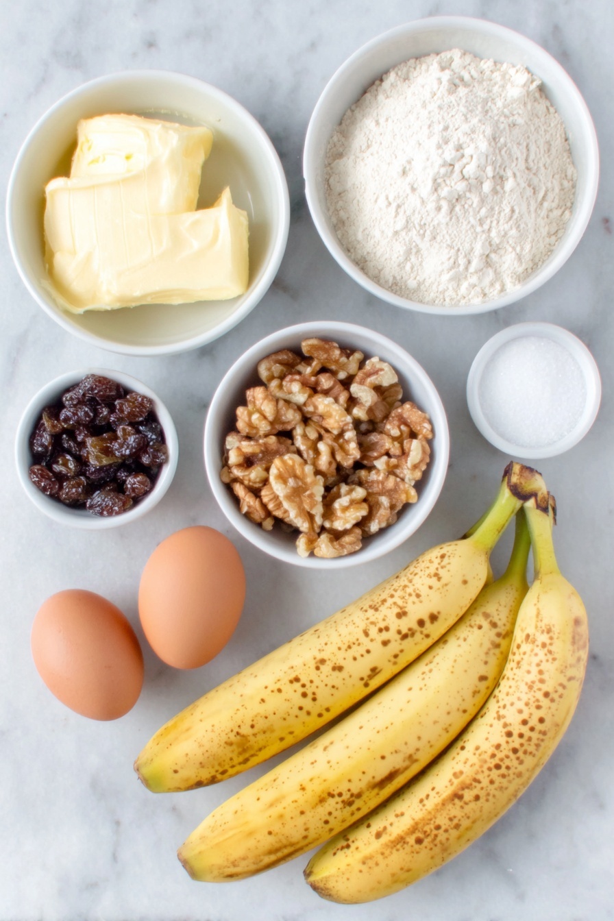 Flat lay of three very ripe bananas with brown speckles, a small white ceramic bowl filled with softened unsalted butter, another white bowl containing granulated sugar, two whole uncracked brown eggs, a white bowl with all-purpose flour, a small white bowl holding walnuts roughly chopped, a small white bowl filled with dark raisins, a small white bowl with pale beige baking soda powder, a tiny white bowl with coarse salt, and a small white bowl with golden vanilla extract, all arranged with perfect symmetry on a clean white marble surface, soft natural light, photo taken with an iPhone, professional food photography style, fresh ingredients, white ceramic bowls, no bottles, no duplicates, no utensils, no packaging --ar 2:3 --v 7 --p m7354615311229779997 - Best Banana Bread, moist banana bread recipe, easy banana bread, homemade banana bread, banana bread with walnuts and raisins