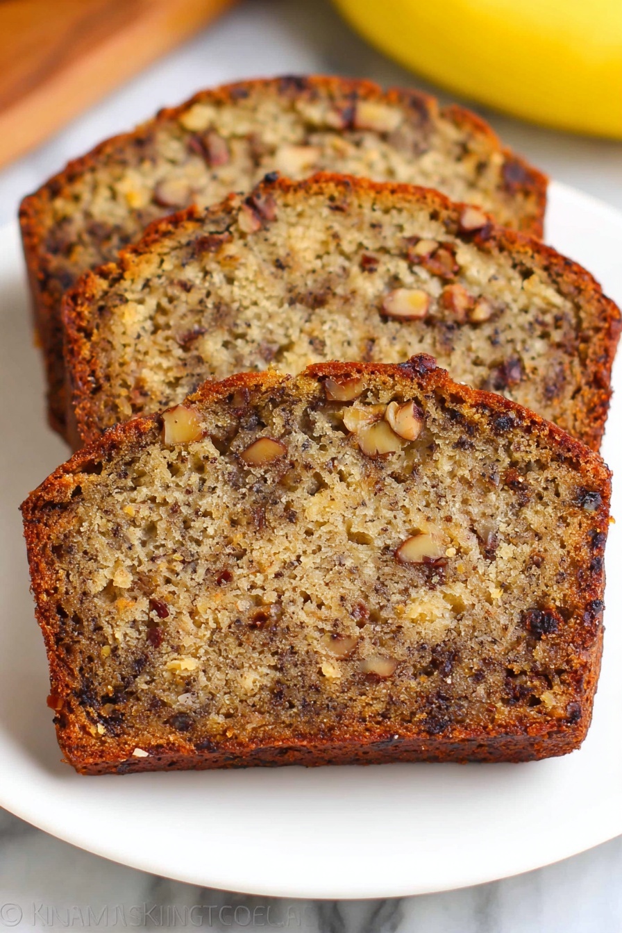 Three slices of banana nut bread are stacked slightly overlapping on a white plate, showing a dense texture filled with dark brown nuts and small chunks of banana inside the golden brown cake. The crust is a darker shade of brown and slightly crisp, while the inside looks moist with a mix of light and dark brown colors from the bananas and nuts. The plate is on a white marbled surface with a blurred yellow banana visible in the background. Photo taken with an iphone --ar 2:3 --v 7 - Best Banana Bread, moist banana bread recipe, easy banana bread, homemade banana bread, banana bread with walnuts and raisins