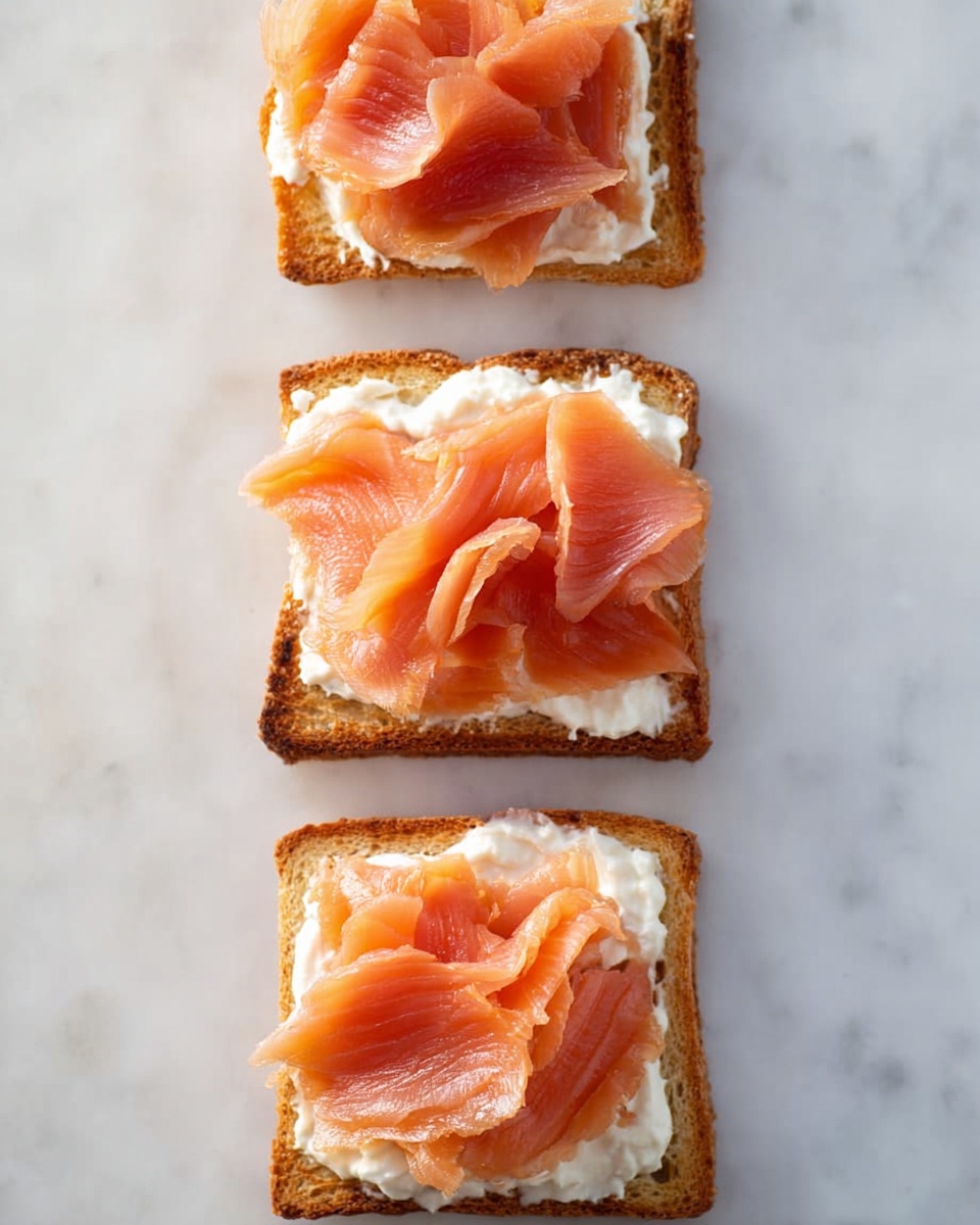The image shows three small square pieces of toasted bread placed in a vertical line on a white marbled surface. Each piece of toast has a layer of white creamy spread topped with a soft, thin slice of orange-pink smoked salmon folded loosely. The toasted bread appears golden brown and slightly crispy on the edges. The arrangement is simple and clean, highlighting the contrast between the creamy white spread and the vibrant salmon color. The photo taken with an iphone --ar 4:5 --v 7