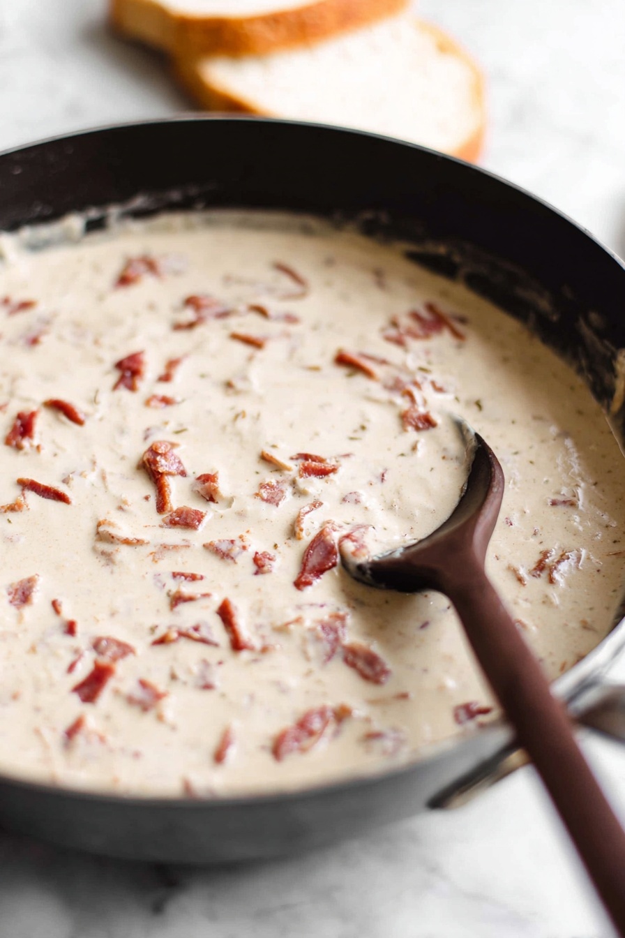 A white plate with ridged edges holds two slices of golden toasted bread stacked slightly off-center, covered generously with a thick, creamy white sauce that has visible small chunks of reddish-brown bacon and tiny bits of green herbs sprinkled on top. The sauce drips slightly over the edges of the toast, and a few small crumbs and herbs are scattered around the plate. The plate rests on a white marbled surface. photo taken with an iphone --ar 2:3 --v 7 - Chipped Beef on Toast, easy chipped beef recipe, hearty breakfast ideas, comfort food recipes, quick savory breakfast