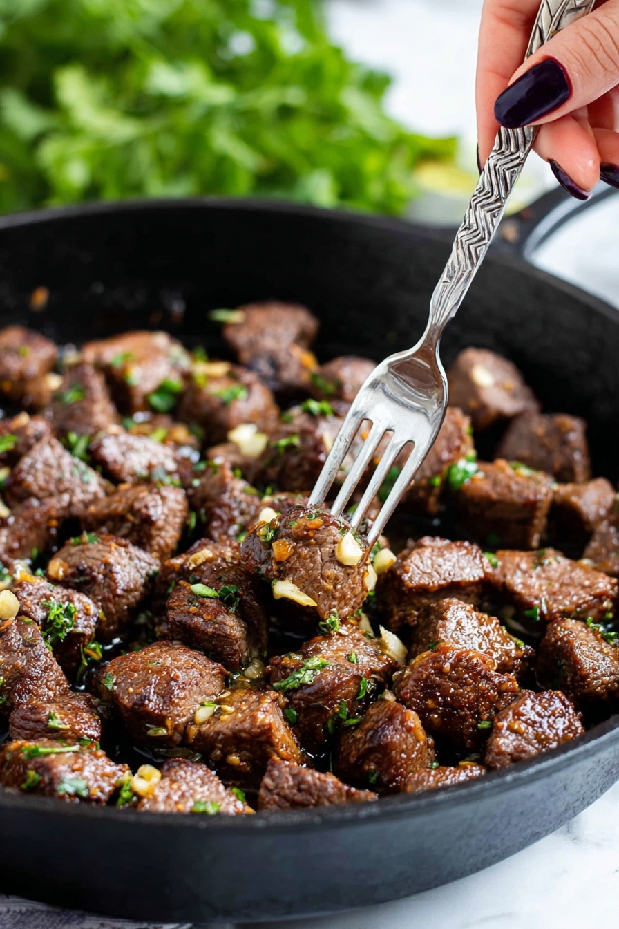 The image shows a black cast iron pan filled with many small chunks of cooked brown meat, each piece sprinkled with bits of green herbs and small pieces of golden-brown garlic. A woman's hand with dark nail polish is holding a silver fork that picks up a single piece of meat from the pan. The background is blurry with green leaves visible, creating a fresh feel. The pan sits on a white marbled surface. photo taken with an iphone --ar 2:3 --v 7 - Garlic Butter Steak Bites, garlic butter steak, steak bites recipe, quick steak dinner, flavorful steak snacks