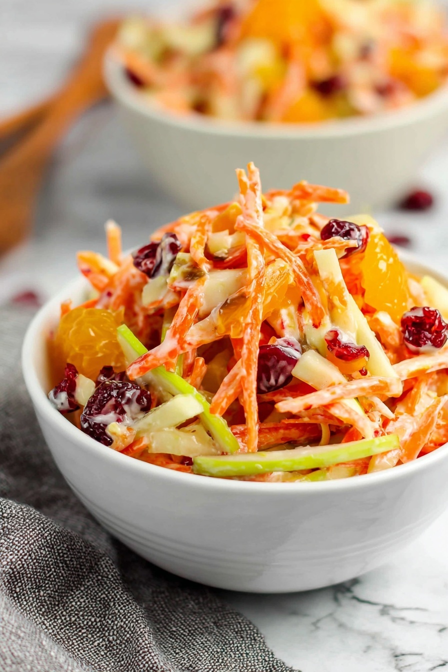 A white bowl is filled with a colorful salad that has shredded orange carrots, thin slices of green apple, small chunks of white apple, bright orange mandarin segments, and dark red dried cranberries mixed together. The salad looks creamy with a light dressing coating the pieces. The bowl sits on a white marbled surface with a gray cloth slightly blurred in the background. Another bowl with the same salad is partly visible behind the main bowl. photo taken with an iphone --ar 2:3 --v 7 - Carrot Apple Salad with Cranberries, healthy fruit salad, easy apple carrot salad, quick summer salads, tangy fruit salad