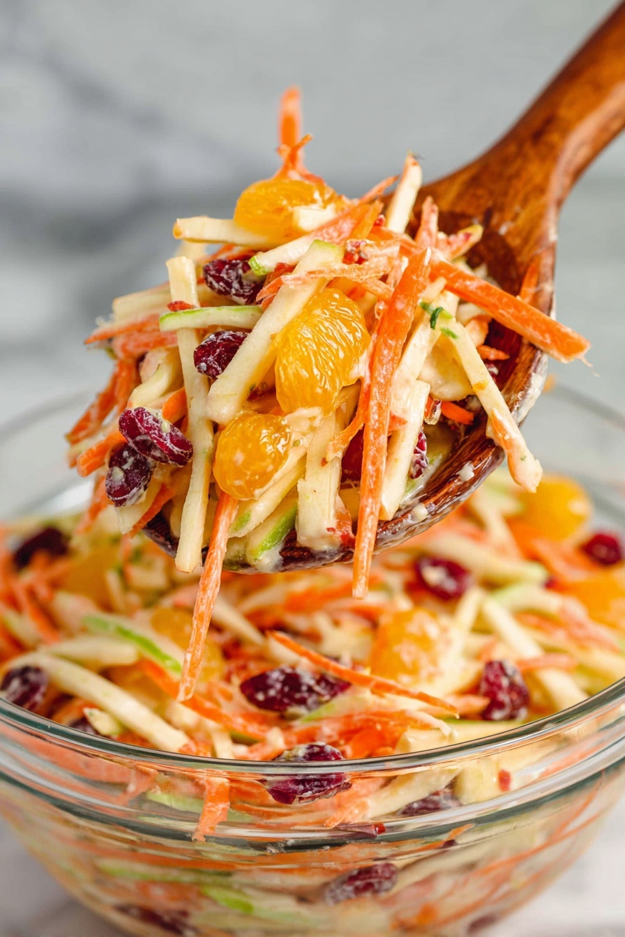 A wooden spoon holds a bright mix of thin, long strips of orange carrot, light beige apple, green apple peels, dark red dried cranberries, and small segments of bright orange mandarin, all coated lightly with a creamy dressing. The spoon is lifted above a clear glass bowl filled with more of the colorful layered salad showing the same ingredients. The background is a white marbled surface, enhancing the fresh colors of the salad, photo taken with an iphone --ar 2:3 --v 7 - Carrot Apple Salad with Cranberries, healthy fruit salad, easy apple carrot salad, quick summer salads, tangy fruit salad