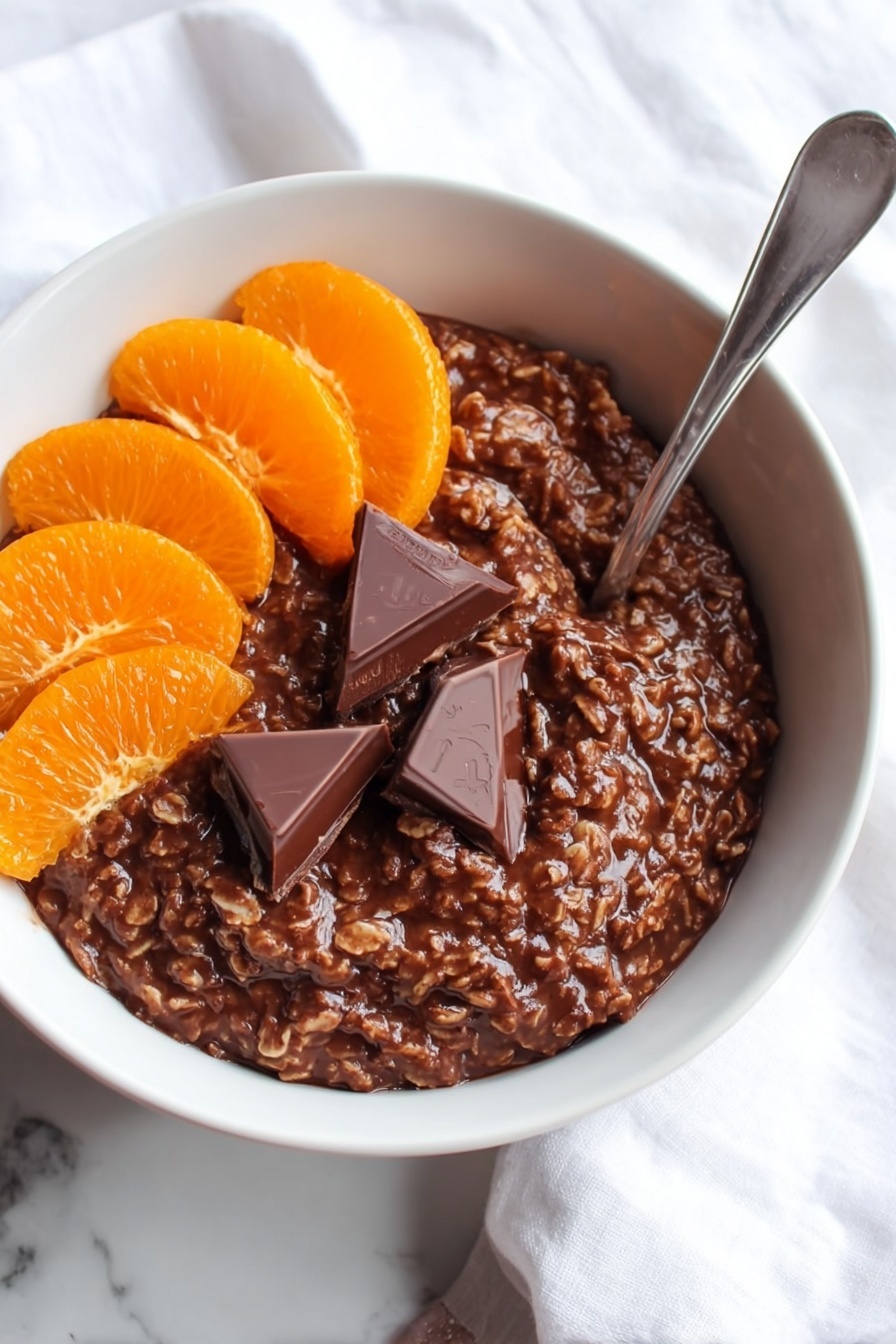 The image shows a white bowl filled with a thick, dark brown chocolate oatmeal mixture that has a slightly rough texture from the oats. On the top left side of the oatmeal, there are six orange slices arranged in a neat cluster, while on the top right side, two triangular pieces of rich, dark chocolate rest partially melted on the oatmeal's surface. A silver spoon is placed inside the bowl on the right side, angled upward. The bowl sits on a white marbled surface with part of a white cloth visible in the background. Photo taken with an iphone --ar 2:3 --v 7 - Chocolate Orange Porridge, easy breakfast ideas, healthy citrus breakfast, quick morning recipes, comforting porridge with chocolate