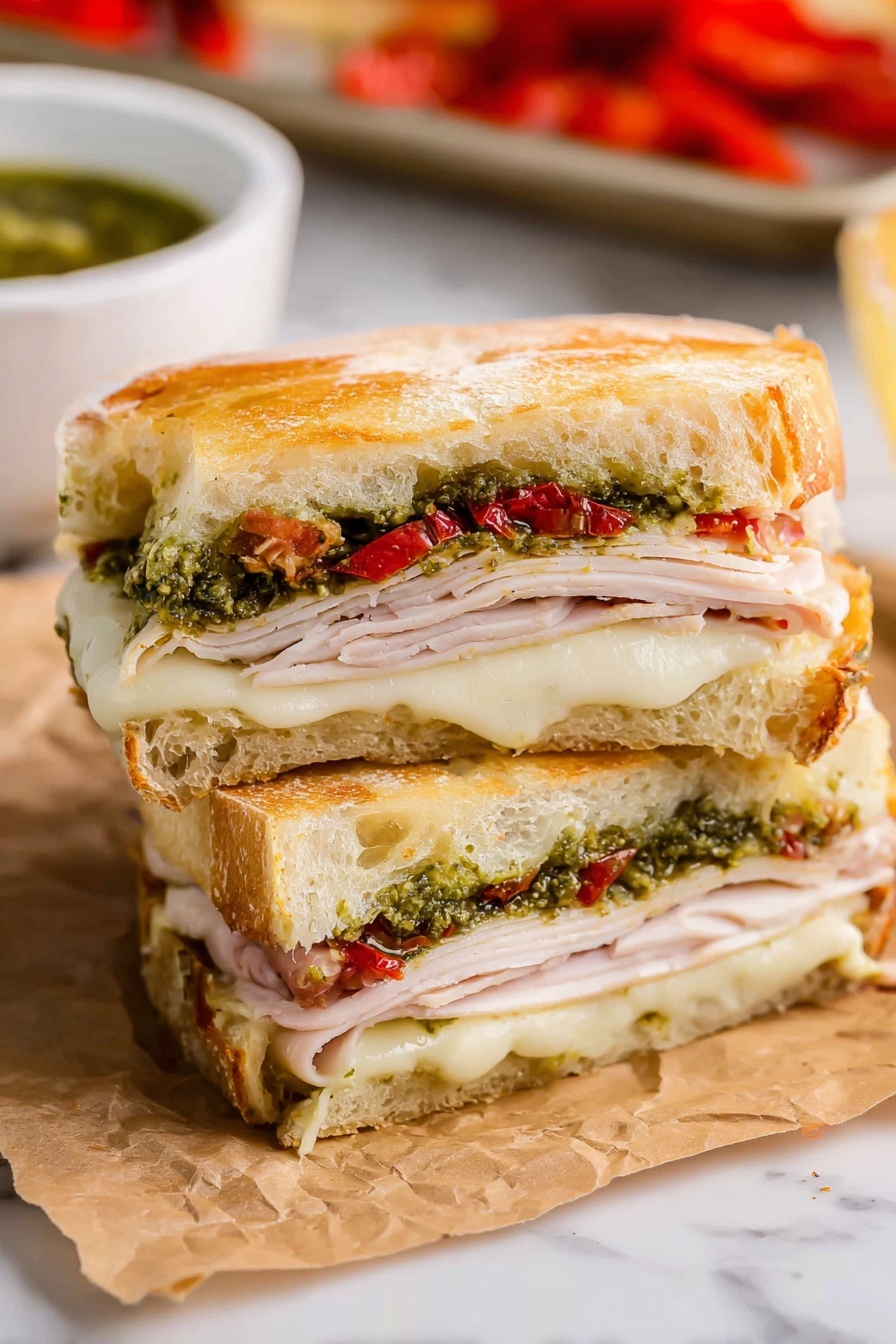 The image shows a sandwich cut in half and stacked, placed on crinkled brown paper against a white marbled surface. The sandwich has three main layers of fillings. The bottom layer consists of melted white cheese with a soft texture. Above it is a layer of thin, light pink slices of turkey. On top of the turkey, there is a green layer of pesto sauce mixed with small pieces of red roasted peppers. The sandwich is held together by two thick slices of golden brown, slightly crusty bread with a soft inside, forming the top and bottom of the sandwich. In the background, there is a white bowl with green sauce and part of a baking tray with more red roasted peppers visible. Photo taken with an iphone --ar 2:3 --v 7 - Copycat Starbucks Turkey Pesto Sandwich, Starbucks turkey pesto sandwich copycat, homemade turkey pesto sandwich, copycat Starbucks sandwich recipe, turkey pesto sandwich at home