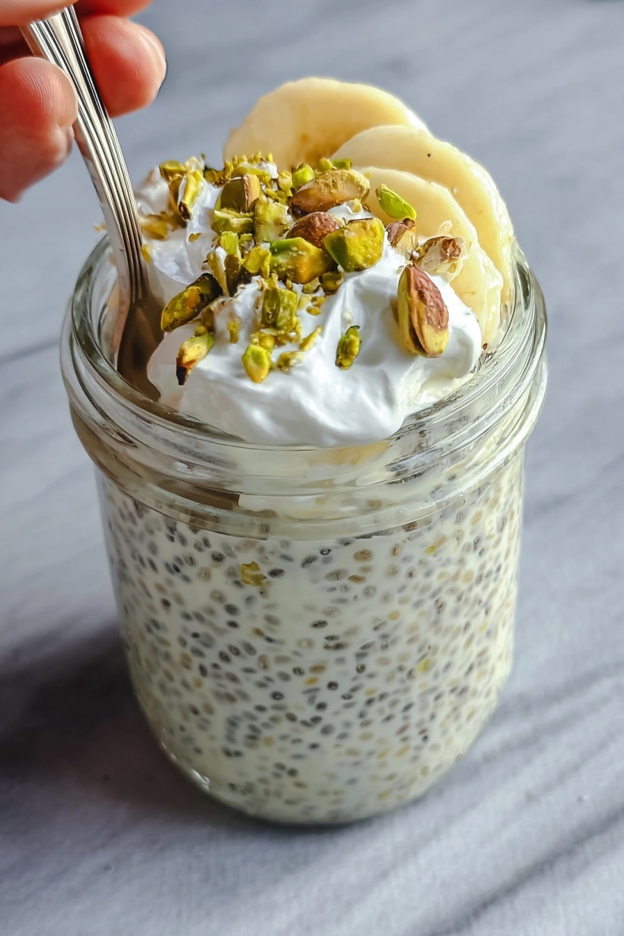 The image shows a clear glass jar filled almost to the top with a creamy white chia pudding that has small black chia seeds spread evenly throughout. On the top layer inside the jar, there are a few slices of light yellow banana, which are partially covered by the creamy pudding. Over the bananas, there is a layer of small white whipped cream dollops, adding a soft texture. Scattered on the very top are roughly chopped green pistachio nuts that add a contrasting color and crunchy texture. A spoon is placed inside the jar, with a woman's hand holding it gently from the left side. The jar is set against a smooth, white marbled surface. Photo taken with an iphone --ar 2:3 --v 7 - Creamy Pistachio Protein Overnight Oats, healthy overnight oats with pistachio, high protein breakfast ideas, make-ahead breakfast recipes, nutritious breakfast overnight oats