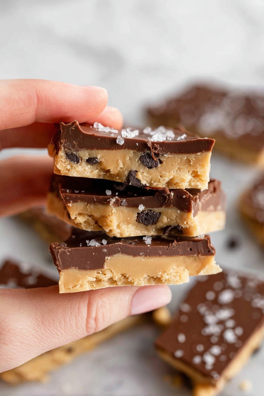 A woman's hand is holding three pieces of snack bark stacked closely together. Each piece has two layers: a bottom light tan layer with a soft texture and dark chocolate chip bits mixed in, and a thick top layer of smooth, dark brown chocolate. The top chocolate layer is sprinkled with small flakes of white sea salt. More broken pieces of the same snack are scattered on a white marbled surface in the background, showing both tan and dark chocolate layers clearly. Photo taken with an iphone --ar 2:3 --v 7 - Healthy Cookie Dough Bark, Healthy Cookie Dough Bark Recipe, Guilt-Free Cookie Dessert, No-Bake Healthy Treat, Easy Healthy Snack