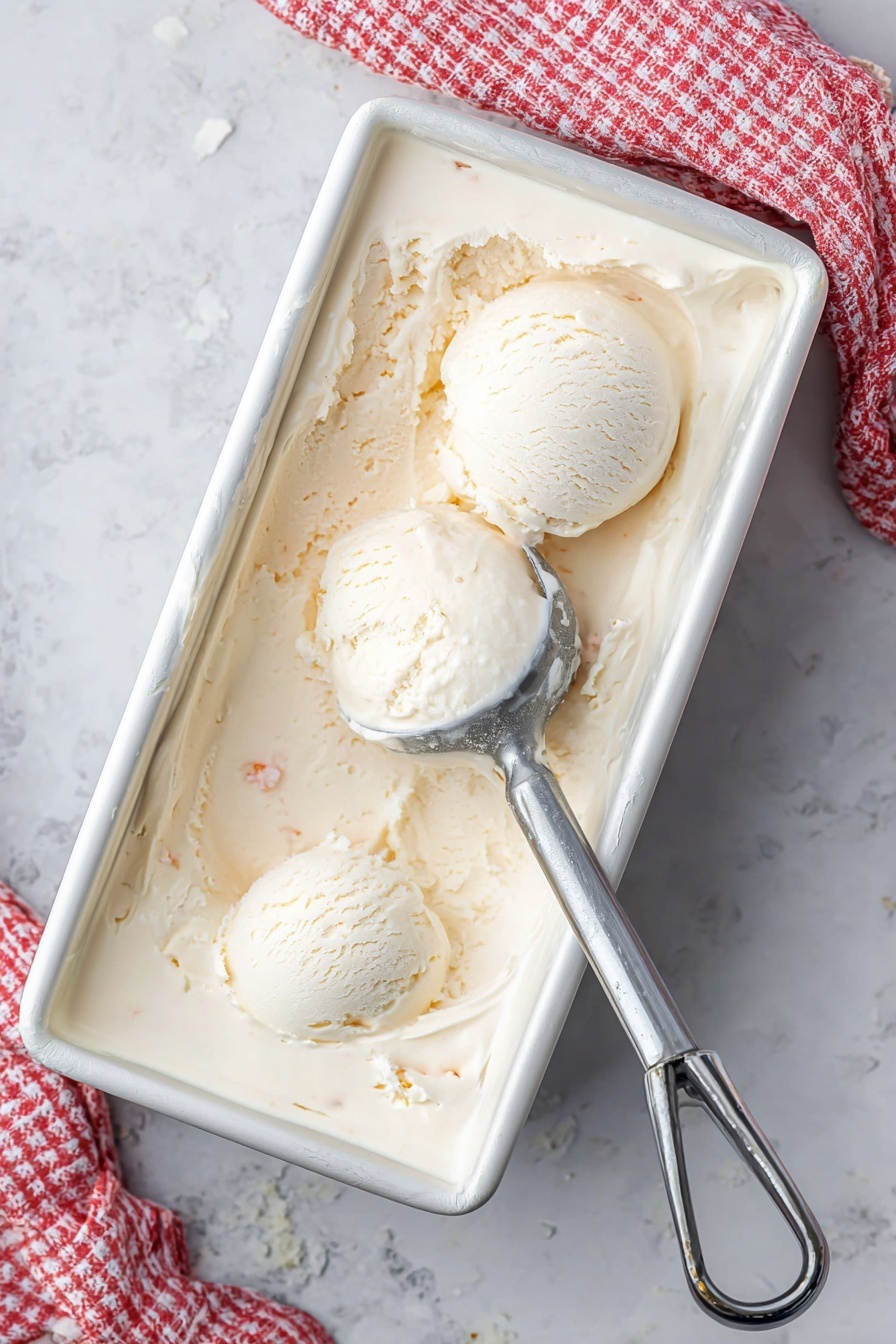 A white metal rectangular container filled with creamy, pale beige ice cream shows two round scoops placed near the top left corner. Ice cream has a smooth but slightly textured surface with small visible bits inside. A silver ice cream scooper rests partially buried on the right side, with some ice cream residue on it. A white and red checkered cloth is partly visible in the upper right corner on a white marbled surface. photo taken with an iphone --ar 2:3 --v 7 - Homemade Coconut Ice Cream, coconut ice cream recipe, tropical frozen dessert, easy coconut ice cream, no-churn coconut ice cream